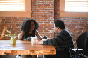 Two disabled Black women (one wearing compression gloves, and the other in a power wheelchair that's partially in view) sit across each other and laugh while drinking coffee.