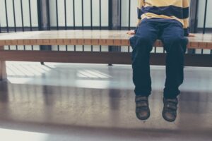 Photo of boy sitting on bench alone - only his lower body and legs are in the picture. He wears a yellow and black striped shirt, black pants and black shoes.