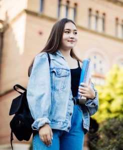 College student standing outside a building holding a backpack and several books