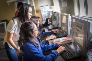 A student sitting and working at a computer as a teacher watches from behind them.