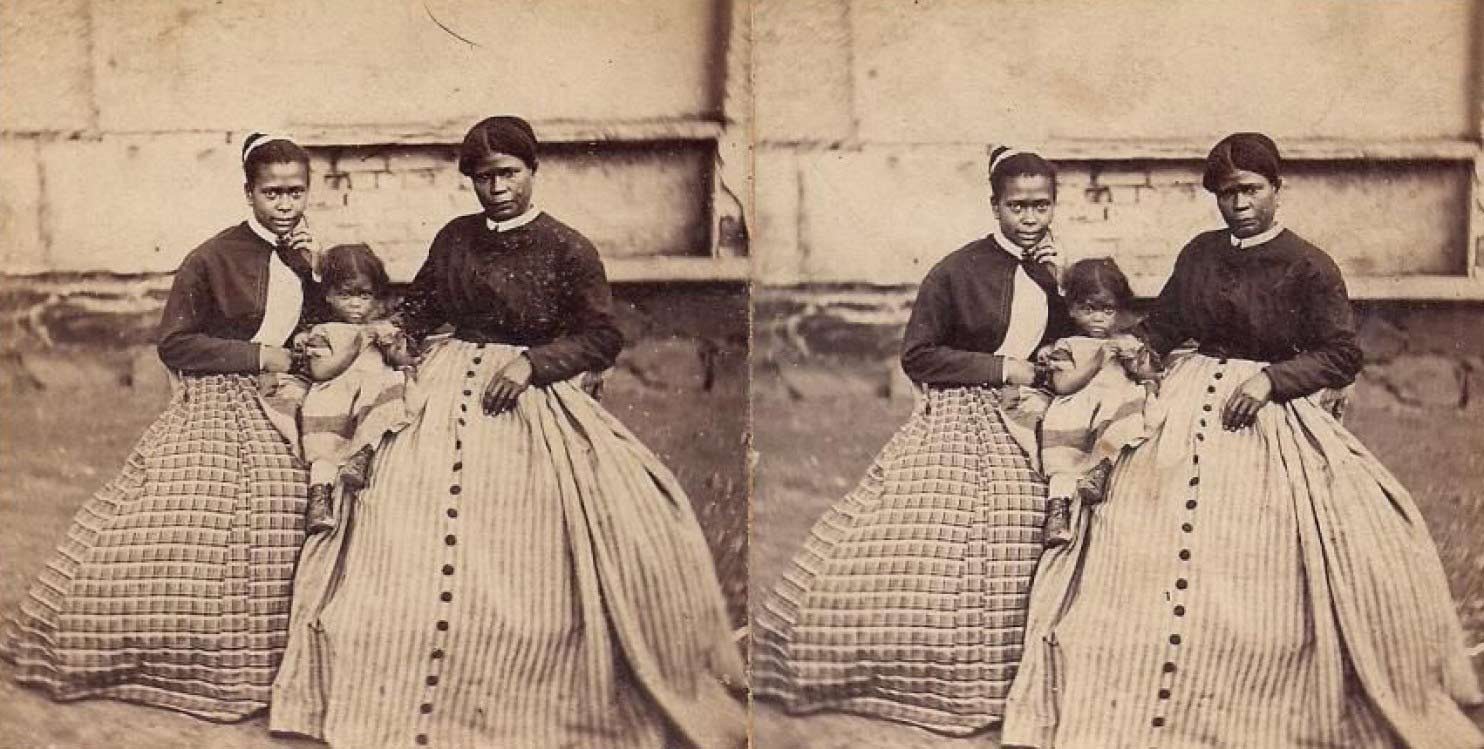 Old photograph of a woman with a teenage and toddler daughter. They are seated, wearing full dresses.