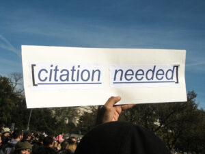 Person holding up a sign that says "Citation Needed"