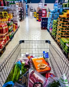 A shoppers-eye view of a grocery cart filled with food in the aisle of a grocery store.