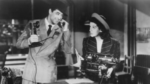 Black and white film screenshot. Cary Grant is on the phone, while Rosalind Russell stands over an old typewriter