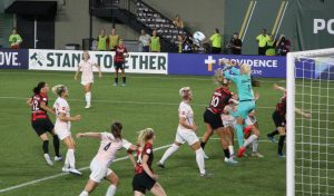 A women's professional soccer match. The ball is in the air and the goalkeeper is lunging for it. Players are crowded around the ball.