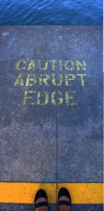 Lettering on a sidewalk says"Caution abrupt edge." The sidewalk ends in water.