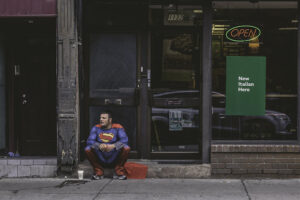 A man in a superman costume sitting in front of a Subway restaurant with a sign in the window advertising the New Italian Hero