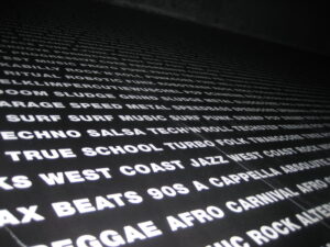 A list of music genres printed on a wall in black and white including Speed Metal Sludge Metal Surf Music Swamp Pop Salsa Turbo Folk West Coast Jazz 90s A Cappella and so on