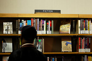 A man at the library browsing a shelf. There is a sign on the shelf that says Fiction.