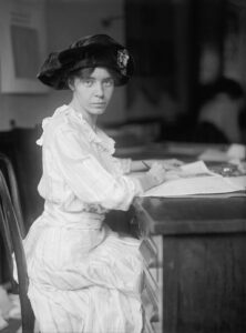 Alice Paul in 1915, sitting at a desk working on some papers. She is wearing a white dress, has her hair pulled back and under a hat, looking rather serious.