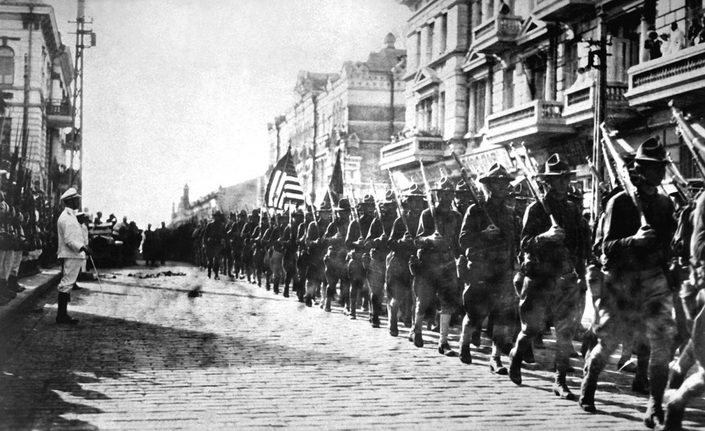 While still fighting in WWI, President Wilson sent American troops to Siberia during the Russian Civil War for reasons both diplomatic and military. This photograph shows American soldiers in Vladivostok parading before the building occupied by the staff of the Czecho-Slovaks (those opposing the Bolsheviks). To the left, Japanese marines stand to attention as the American troops march.