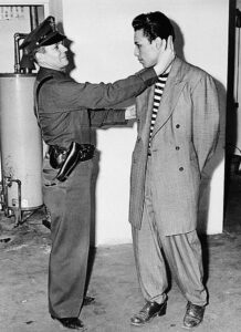 A young Mexican-American man in an oversized suit being arrested by an officer. 