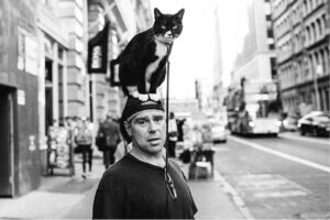 Black and white photo of a man standing on a sidewalk with a cat sitting on his head.