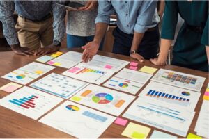 A table with lots of different graphs and charts spread out on top of it. There are people standing nearby examining them.