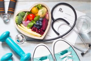 A collection of things, including some fresh vegetables arranged in a heart dish, a stethoscope, some weights, a cup, and a prescription pad.