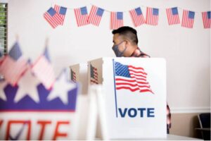 A person in front of a podium that reads "Vote" in front of an American flag. There are several other American flags in the room.