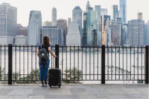 A woman standing with a suitcase looking out over a river in front of a city.