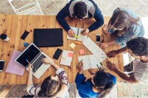 Several people gathered around a table with papers, a computer, and other work materials.