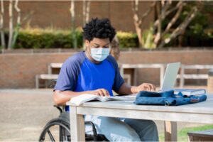A person in a wheelchair and wearing a disposable mask sitting at an outdoor table working on a laptop.