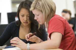 two students sit side by side, actively looking at learning material