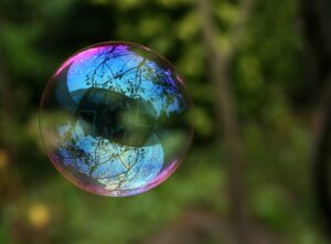 A close-up of a soap bubble floating in front of trees with nearby building and sky reflected in the bubble surface