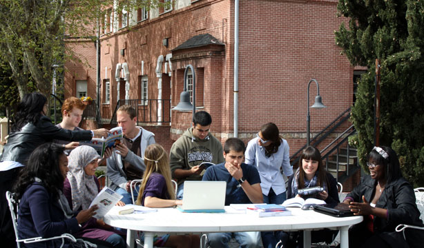 Diverse group of students outside at a table communicating and interacting. Laptop computer is open on the table as well as books and notebooks.
