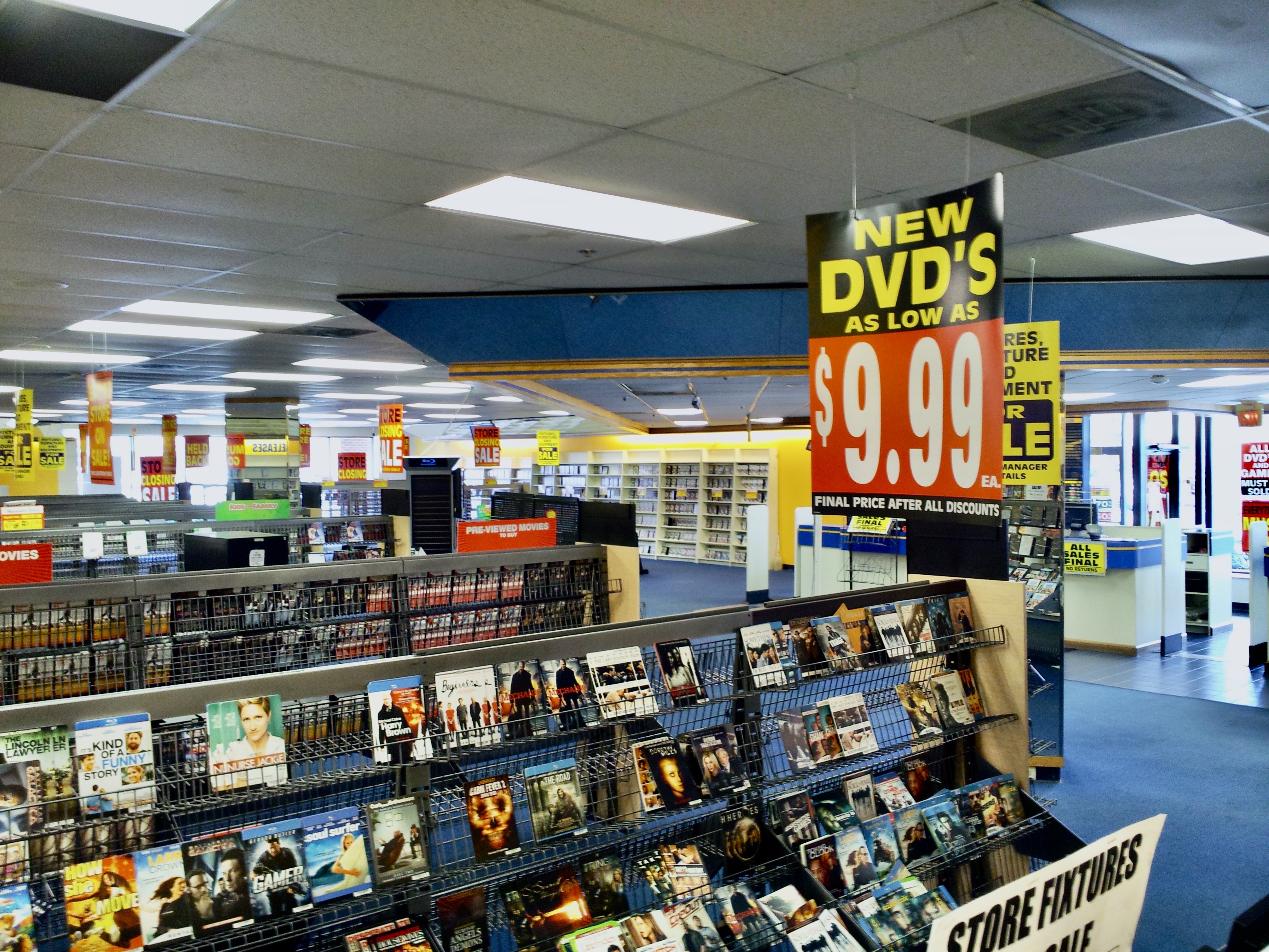 Inside of a Blockbuster as it's going out of business. Shows signs advertising new DVDs as low as 9.99 and shows displays of DVDs and videos.