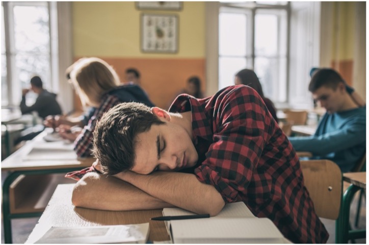 A student asleep at their desk amidst other students taking notes.