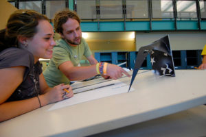 Man wearing green shirt and woven bracelets helping female student with architecture project on a drafting table