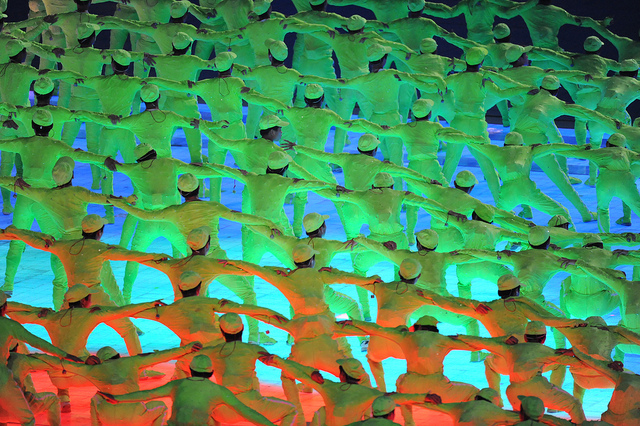 Lines of dancers in an outdoor performance, arms placed around one another's shoulders. They are backlit in waves of orange, green, and blue