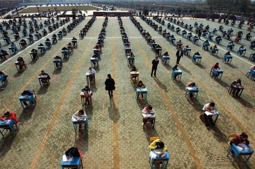 rows of students in desks outdoors, taking an exam