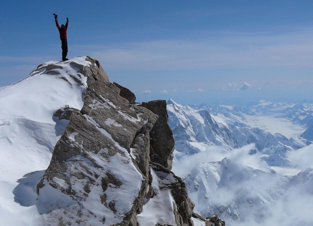 A hiker at the top of a mountain. Other mountain peaks can be seen at lower elevations. The hiker is raising their hands in triumph.