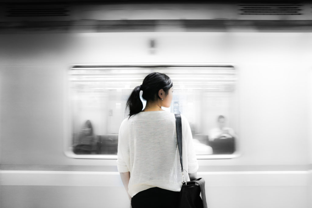 black and white photo of a woman standing in front of a subway as it zips past her.