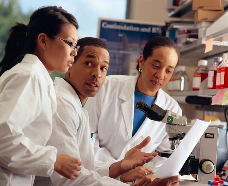 Three researchers review data while talking around a microscope.