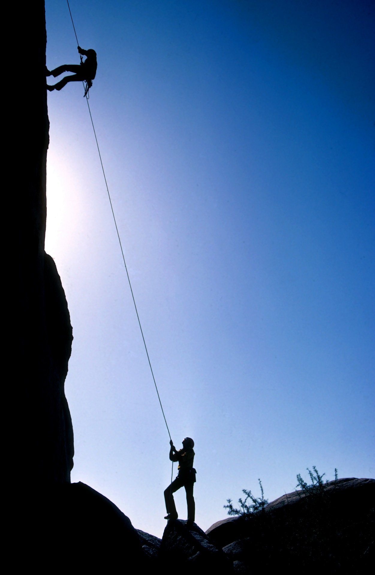 Photo of a man rappeling down a cliff-face while someone belays for him, holding the rope down on the ground. 