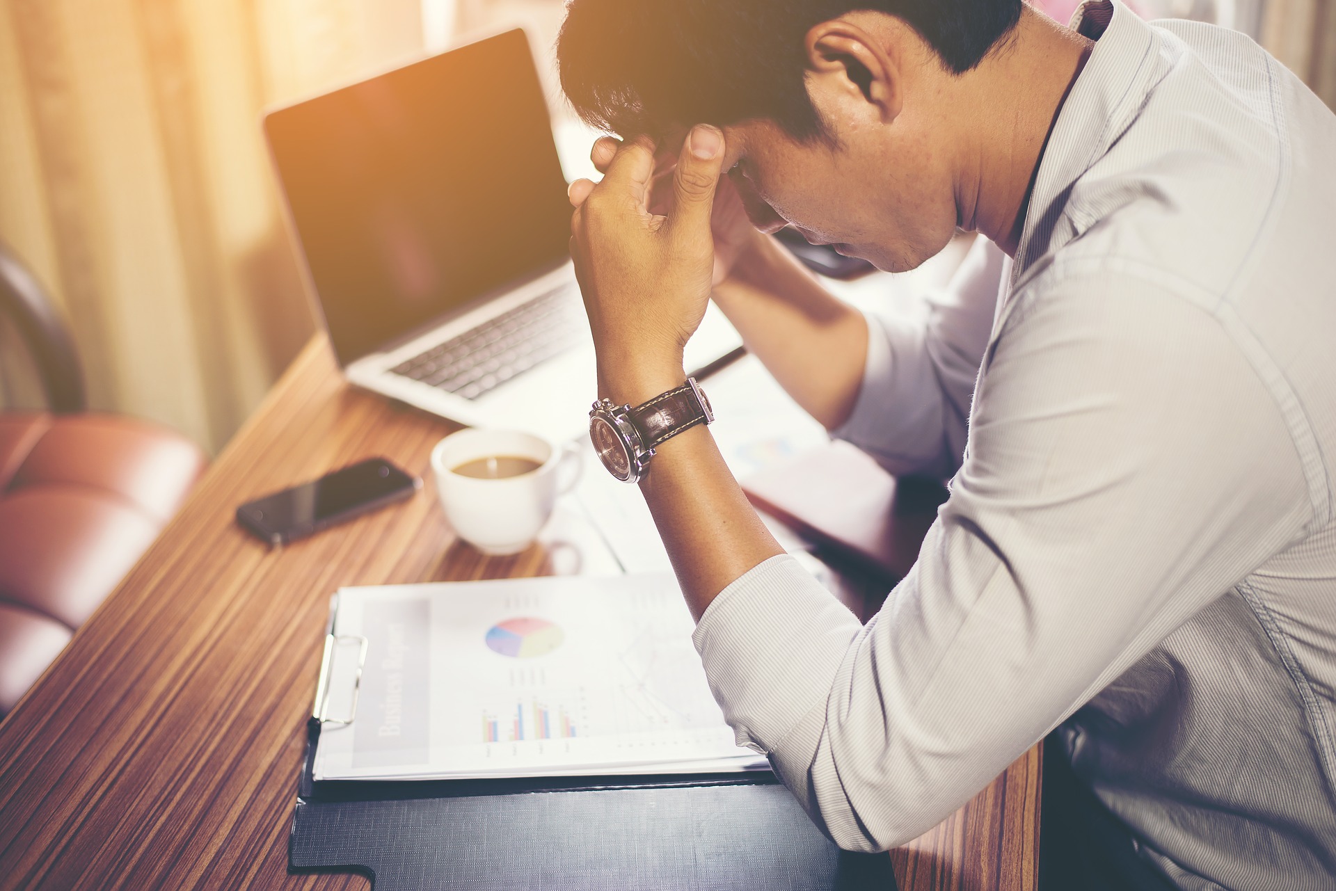 Man sitting at a desk with his laptop and a coffee, looking over some work papers with his hand on his head, appearing to be stressed out.