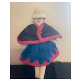  a handmade folk-art doll, wearing traditional-style clothing including a woven hat, a dark cape wit