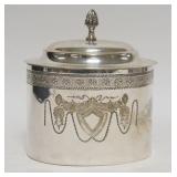 1201	SILVER PLATED OVAL COVERED JAR HAS A SHIELD & FLOWER DESIGN ON BOTH SIDES. 6 1/2 IN X 5 IN 7 IN