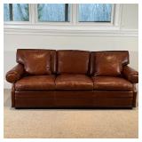 Brown leather sofa, three cushions with two accent pillows, on a lower wood trim [appearing in overa