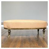 OVAL UPHOLSTERED OTTOMAN WITH BRASS CASTERS | This oval ottoman is upholstered in a light tan textur