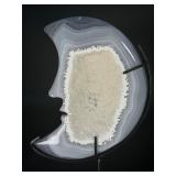 A luminous Botswana Agate druzy carved into a moon shape, showcasing delicate bands of soft gray, caramel, and cream. The sparkling druzy surface catches the light, in a custom metal stand.