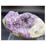 A natural amethyst geode filled with shimmering violet quartz crystals, accented by soft white calcite and encased in its original stone shell.