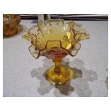 The image displays a collection of vintage amber glass items, including a ruffled compote, a basket dish, a covered candy dish, and a small mug