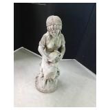 Cement statue of a lady sitting on a tree stump holding a water pitcher with a basket of fruit alongside her