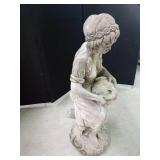 Cement statue of a lady sitting on a tree stump holding a water pitcher with a basket of fruit alongside her