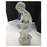 Cement statue of a lady sitting on a tree stump holding a water pitcher with a basket of fruit alongside her
