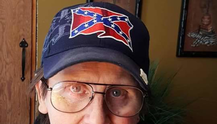 A person wearing glasses and a dark baseball cap embroidered with a Confederate flag.