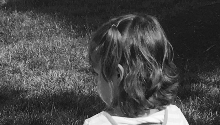 Black and white view from behind a young child with wavy hair and a small pigtail, looking away outdoors on grass.
