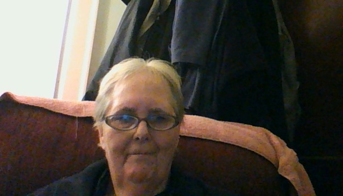 An elderly woman with short white hair and glasses smiling gently while sitting in a chair.