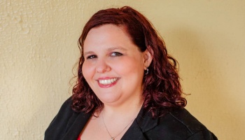 Smiling woman with curly auburn hair wearing a black blazer against a yellow wall.